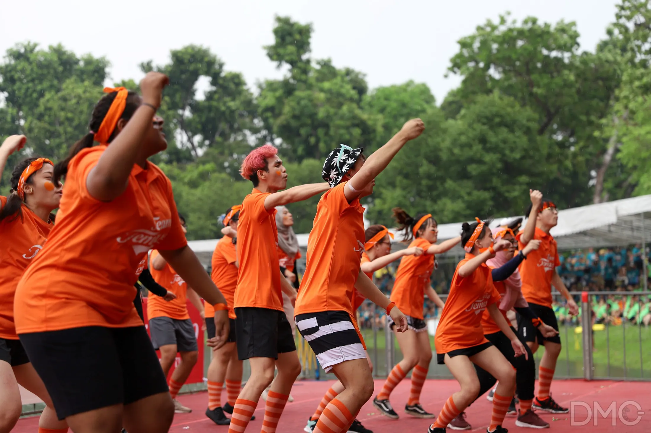 TP students in matching orange outfits cheer energetically during an outdoor event.