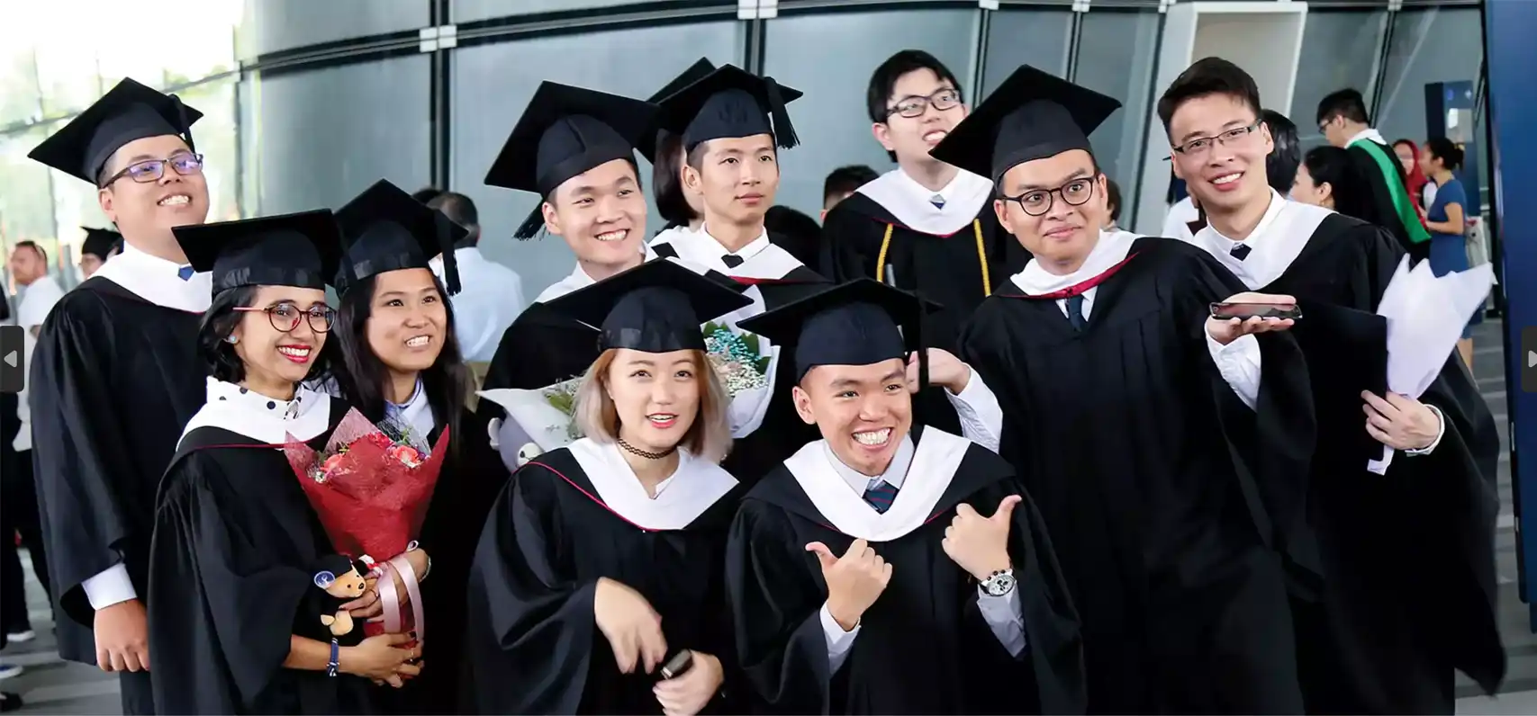 A group of university graduates in black academic gowns and caps celebrating together at a graduation ceremony. They are smiling, posing for a photo, and holding bouquets and certificates.