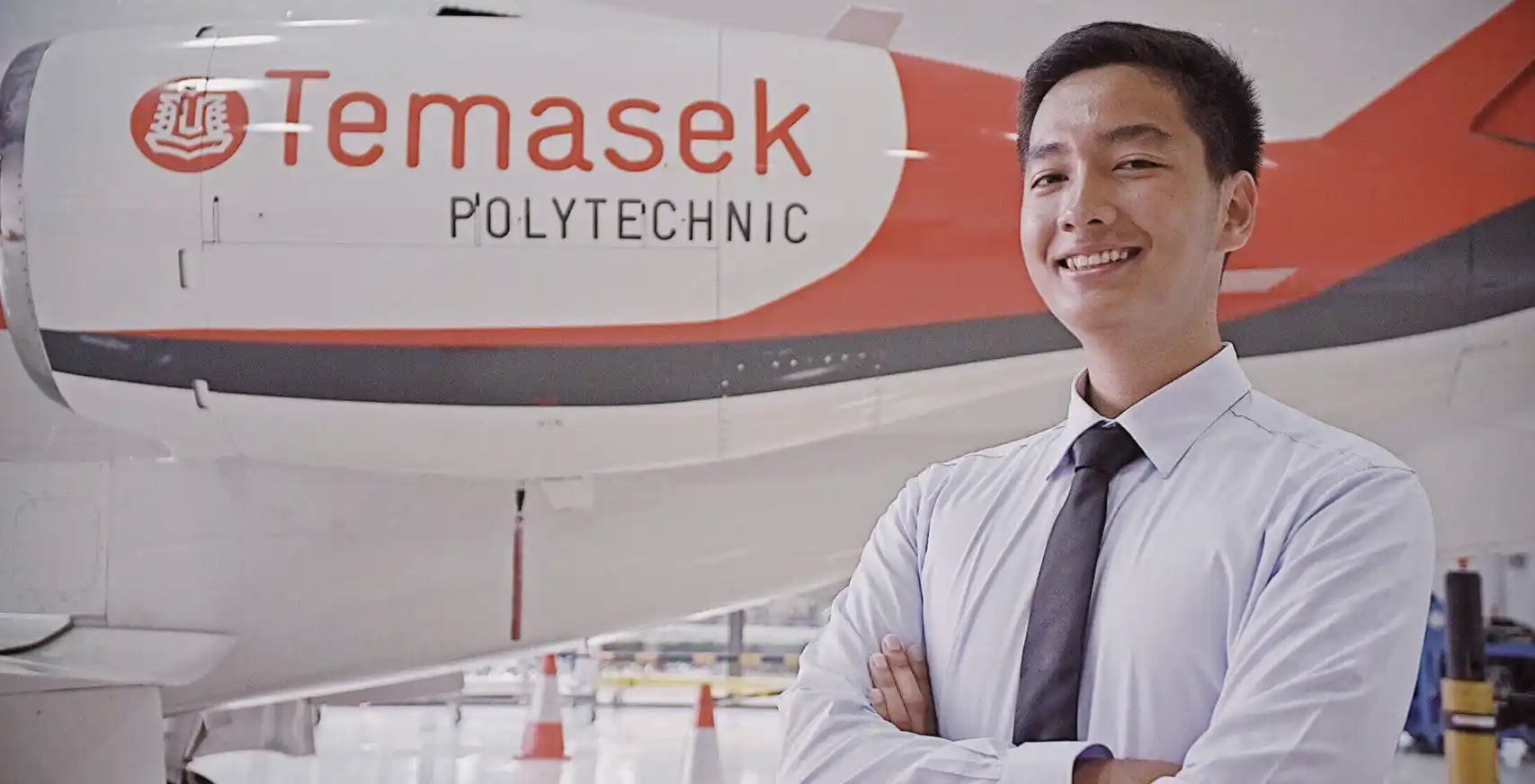A photo of Noorhakim B Jasman, smiling while posing with arms folded in front of a Temasek Polytechnic aeroplane model.