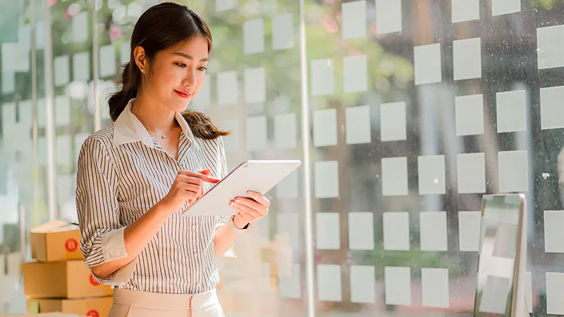 A smiling, focused young professional woman in a striped shirt using a tablet in a modern office with blurred glass walls and boxes in the background, symbolizing digital learning and career development through the SCTP initiative.