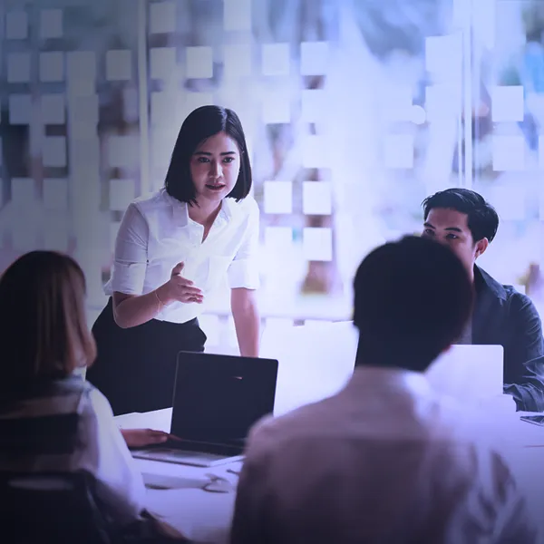 Corporate training session in a modern office with four professionals collaborating around a conference table. One team member stands and leads the discussion, while others listen attentively. Glass wall with colorful sticky notes in the background suggests active brainstorming and project planning.