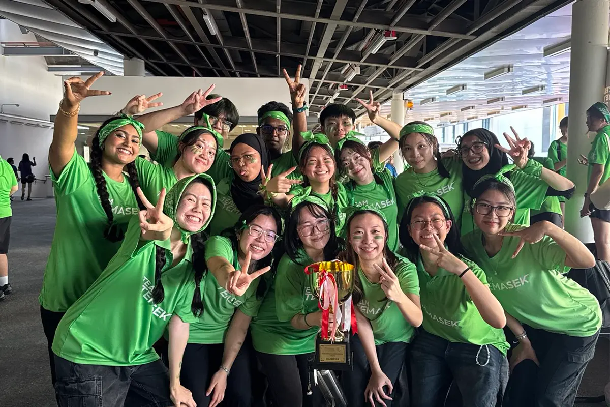 A group of Temasek Polytechnic students in green shirts celebrating with a championship trophy. They are smiling, posing with peace signs, and appear to be at a school sports team event.